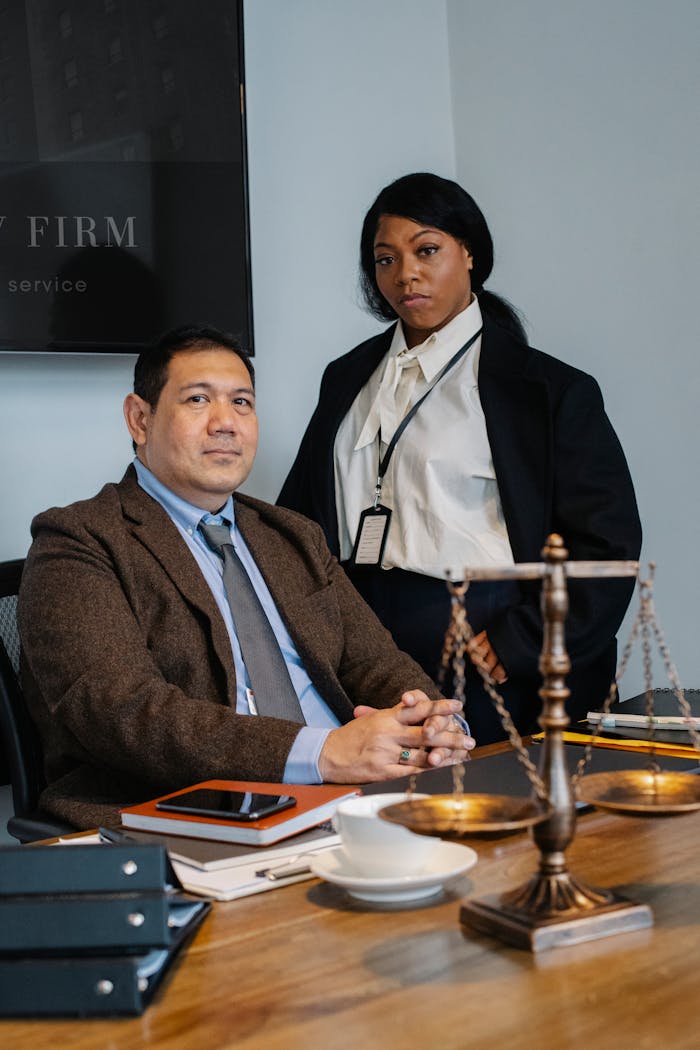 services-03 Focused young black woman in formal clothes standing near male ethnic colleague at wooden table with documents and justice scale during work in law firm