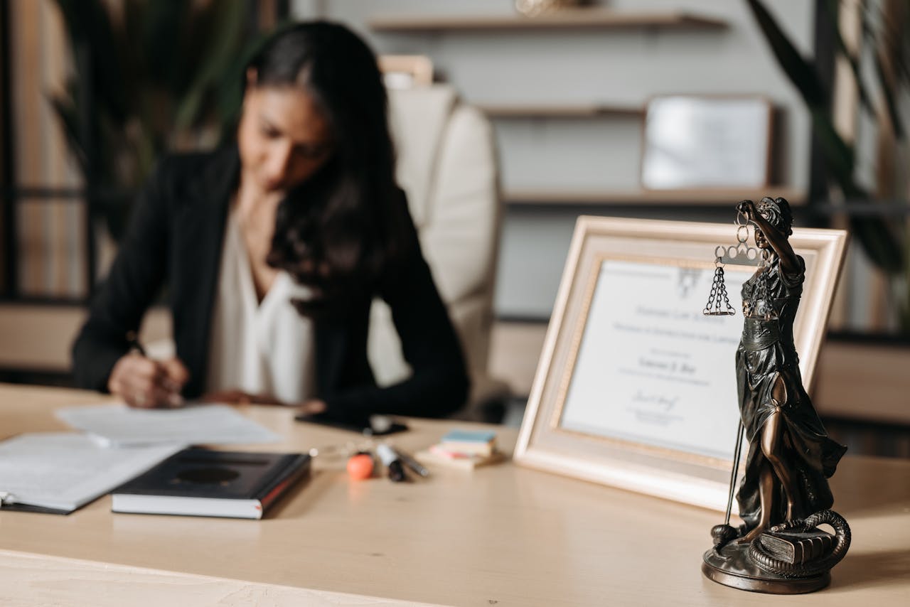 hero-img-01 Focused woman writing at desk with Lady Justice figurine and certificate in office.